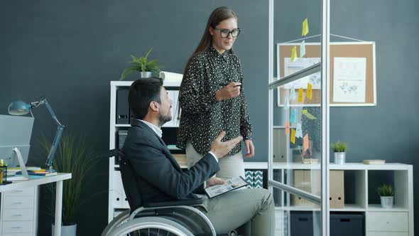 Young Woman and Disabled Man in Wheelchair Talking Writing on Sticky Notes on Glass Board in Office