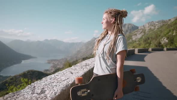 Pretty dreadlocks woman longboarder  enjoying amazing view on mountain holding skateboard