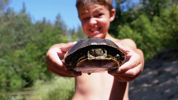 Boy Holds Turtle in Arms and Smiles Viciously on River with Green Vegetation