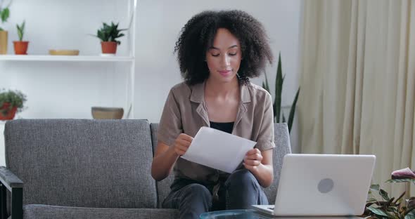 American Woman in Casual Clothes Sits at Home in Office on Sofa with Laptop, Picks Up Envelope From