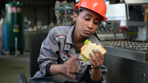 A young black woman in an orange helmet eats a sandwich in a production facility