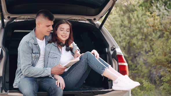 Couple Sitting in Car Trunk and Reading Book Together