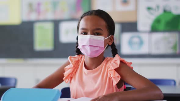 Portrait of mixed race schoolgirl wearing face mask, sitting in classroom looking at camera