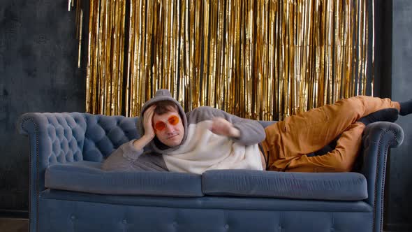 Young Man Corrects Hood with Ears and Bright Glasses Lying on Sofa