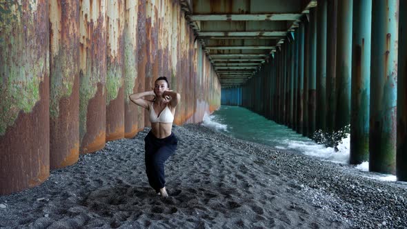 a Barefoot Woman Dances Under the Pillars of the Bridge Against the Background of the Incoming Waves