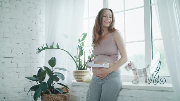 Smiling Pregnant Woman Listening Music with Earhpones at Modern Home.