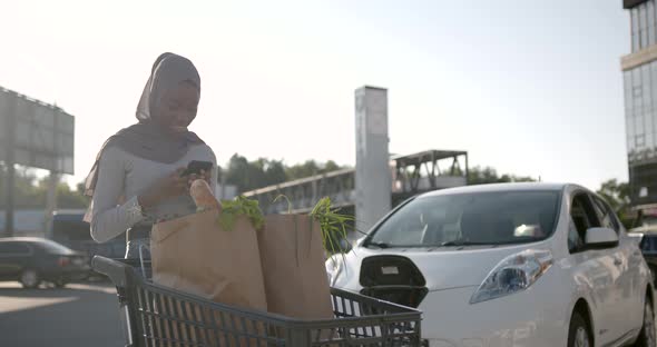 Muslim Young Woman Using Phne While Electric Car is Being Charged