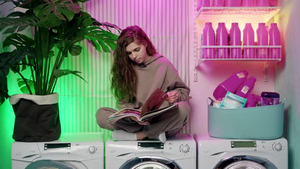 Beautiful Young Woman with Long Brown Wavy Hair in Beige Tracksuit Sits on Washing Machines and