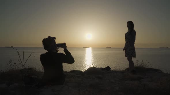 One girl taking picture of another girl with phone in the sea sunset view on the hill