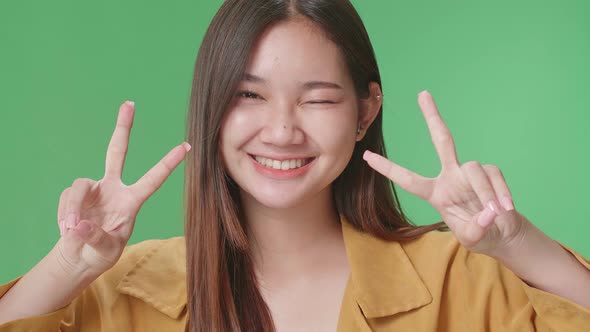 Smiling Young Asian Woman Showing Gesture Peace While Standing On Green Screen In The Studio