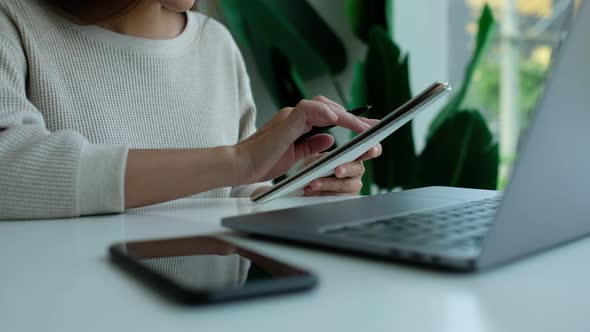 a woman reading and writing on a notebook with laptop computer and smartphone on the table