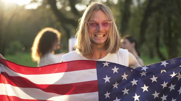 Excited Young Man Posing with American Flag in Sunbeam Outdoors with Blurred Women at Background