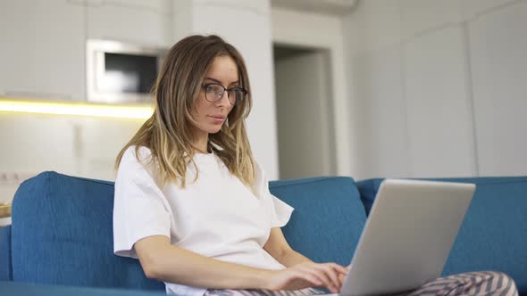Blonde Woman in Pajama Sit on the Couch and Typing on Silver Laptop Low Angle View
