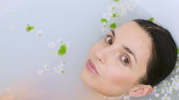 Brunette Girl Lies in a White Bathtub with Milk and Small Flowers Floating