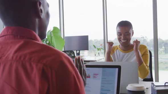 Happy african american creative businesswoman celebrating at desk in office with male colleague