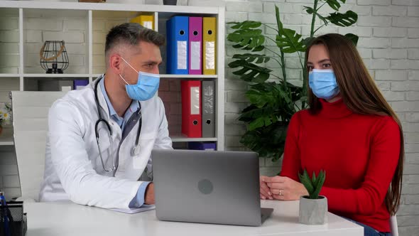 Doctor and patient wear protective masks on faces in hospital office talk listen