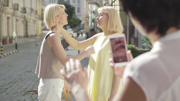 Woman Taking Photo of Female Friends on Smartphone Outdoors. Cheerful Caucasian Mid-adult Tourists