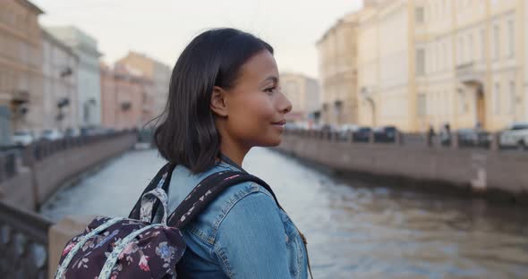 Happy African Female Traveler with Backpack Standing Near River on Old City Street