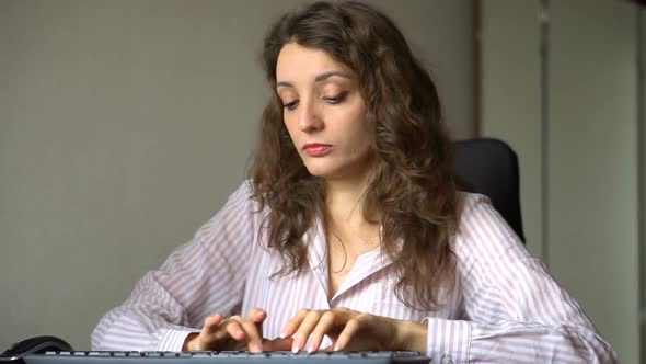 Young Female Office Manager in White Shirt and Curly Hair is Sitting at the Table and Typing Using