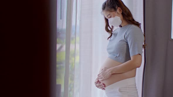 Pregnant Woman wear medical face mask protect virus standing in front of windows stroking big belly