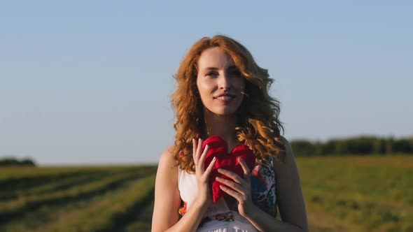 Redhaired Woman in a White Dress Holding a Soft Heart on Background of Field