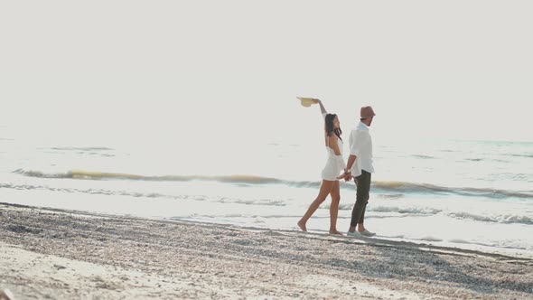 Romantic Couple in Hats Walking Along Sandy Beach Holding Hands