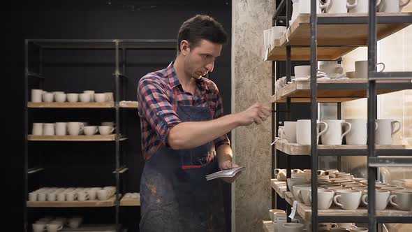 Focused Male Potter Counting Cermics from the Shelves and Noting in the Notebook