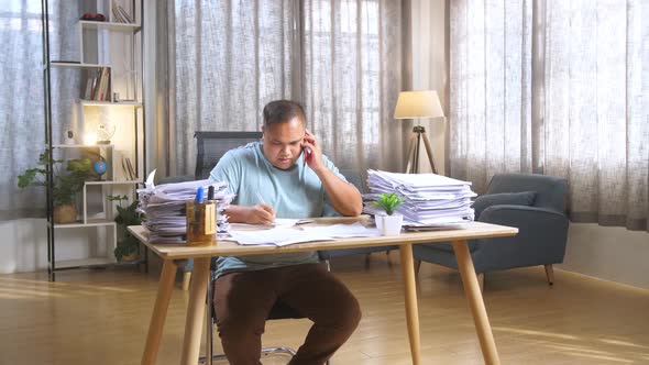 Fat Asian Man Talking On Phone And Taking Note While Working Hard With Documents At Office