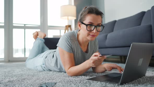 Woman with Glasses is Lying on the Floor and Makes an Online Purchase Using a Credit Card and Laptop