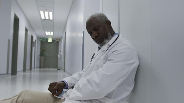 Tired african american male doctor sitting on hospital corridor holding face mask