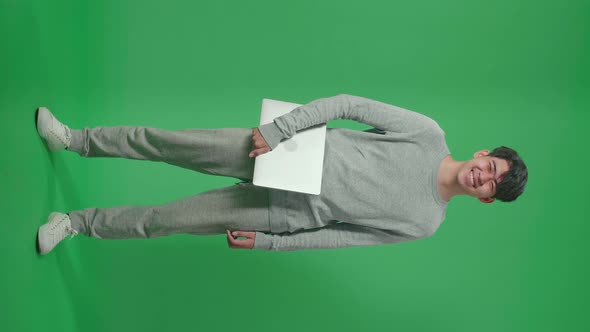 Full Body Of Smiling Asian Man Holding Computer While Standing In Front Of Green Screen Background