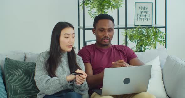 Mixed Ethnicity Couple Sitting on Sofa Purchasing Online Using Credit Card and Laptop Computer