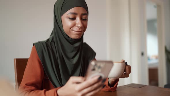 Pretty Muslim woman drinking tea and typing by phone