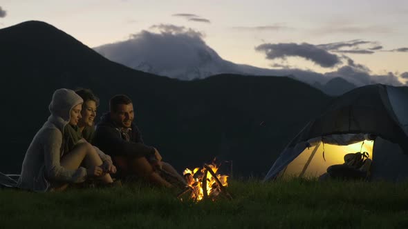 Group of Three Friends Warming with Camp Fire in Nature Mountain Outdoor Camping Scene at Night