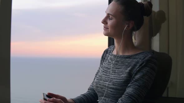 Smiling Girl Standing By the Window During a Sunset and Listening to Music with Headphones Using