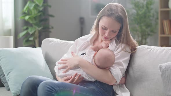 Mom Breastfeeds Her Little Daughter in the Morning Sun in the Living Room