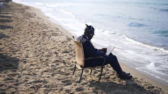 Wide Shot of Relaxed Elegant African American Man Enjoying Music in Headphones Sitting on Wicker