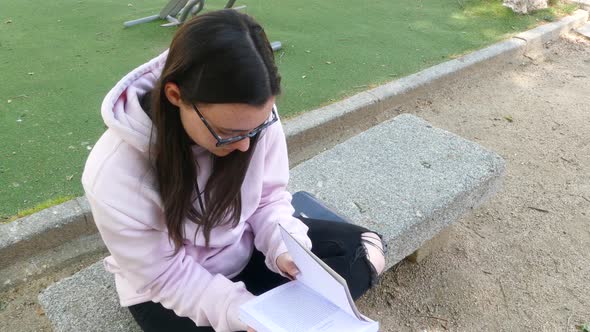 Young and cute girl with glasses reading a novel on a stone bench in a park.