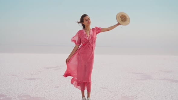 Woman in Blowing Airness Pink Dress and Hat Standing on Salty Beach on Pink Lake