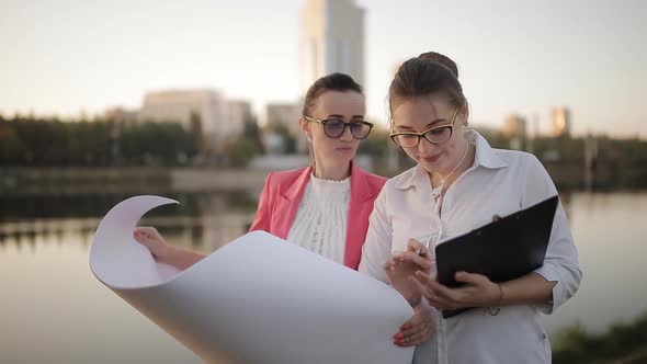 Two Women with the Blueprints in Hand in the City on the Waterfront on the Background Highrise