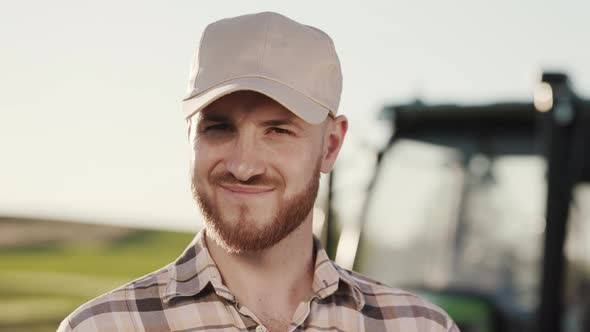 A Portrait of a Young Bearded Farmer is Standing in a Field Near a Tractor