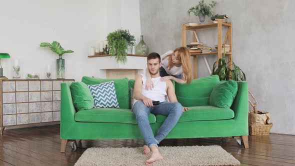 Young Couple in Living Room on Green Sofa, Man Wants To Watch TV, Woman Hugs Him