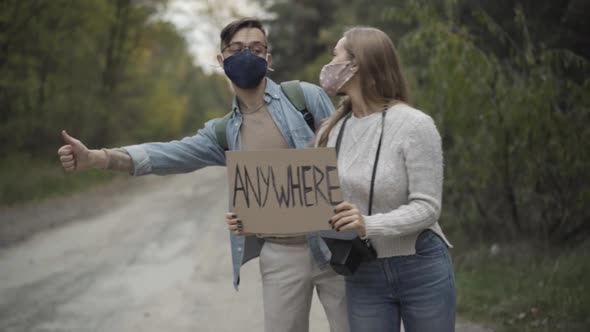 Young Caucasian Boyfriend and Girlfriend in Covid19 Face Masks Hitchhiking with Anywhere Cardboard