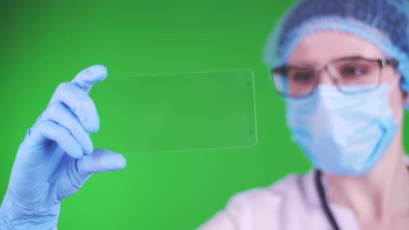 Green Background. Close-up, Doctor Dressed in Medical Cap, Mask, Blue Medical Gloves, Holds a Glass