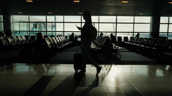 A Woman Is Walking Along the Terminal of the Airport, Carrying a Bag on Wheels. In the Hand