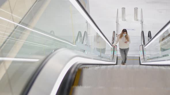 a Young Woman in a Protective Mask Climbs the Escalator and Looks at the Phone
