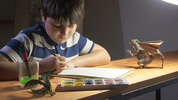 A Boy Draws Watercolor Sitting at Table on Which Stand Toy Dragons