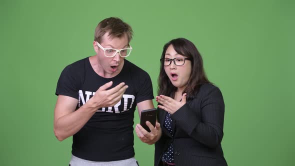 Mature Asian Businesswoman and Young Scandinavian Nerd Man Using Phone While Looking Shocked