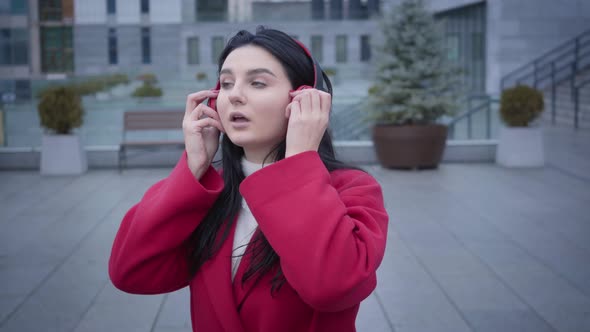 Cute Caucasian Girl with Black Hair and Green Eyes Putting on Headphones and Moving Outdoors. Joyful