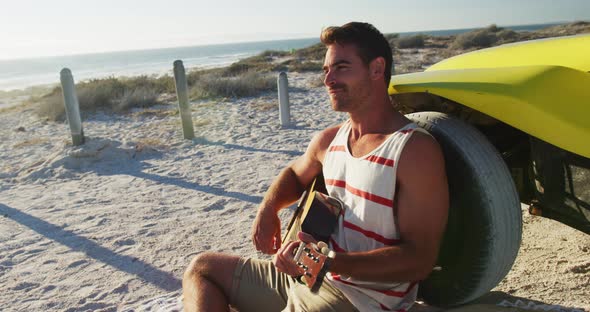 Happy caucasian man sitting beside beach buggy by the sea playing guitar